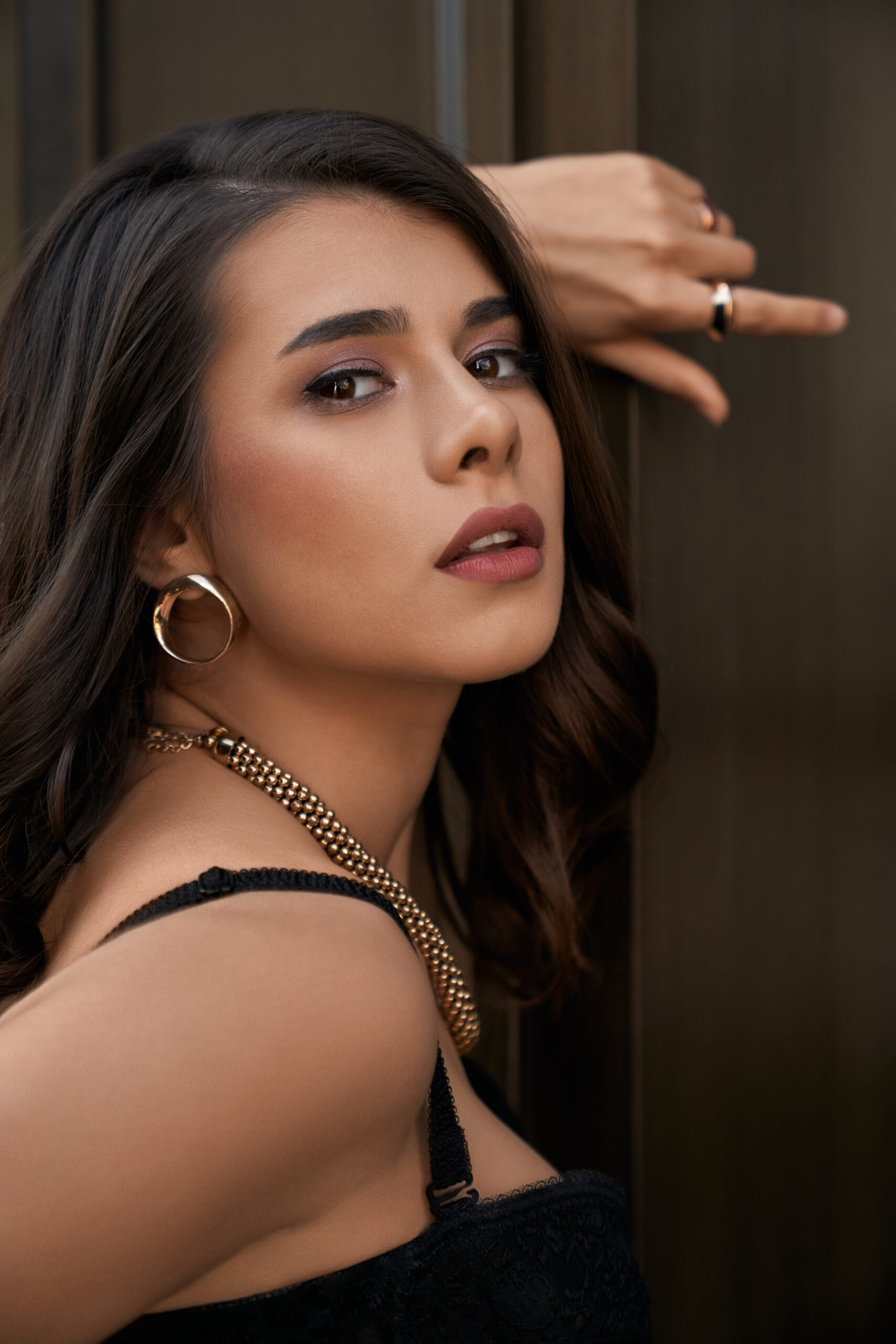 Side view of young caucasian stunning woman wearing black lace corset and golden accessories looking at camera. Closeup portrait of female model with perfect make up posing near wooden door outdoors.